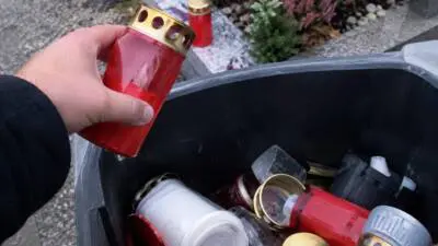 KI-generiertes Bild: Eine Hand hält eine rote Grabkerze über einem Behälter mit gebrauchten Grablichtern.