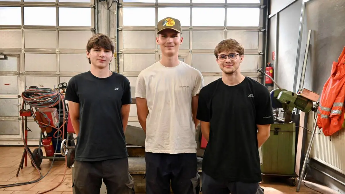 Drei junge Männer stehen in Arbeitskleidung vor Werkstattausrüstung.
