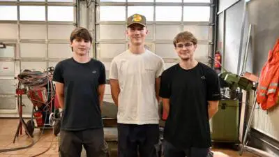 Drei junge Männer stehen in Arbeitskleidung vor Werkstattausrüstung.