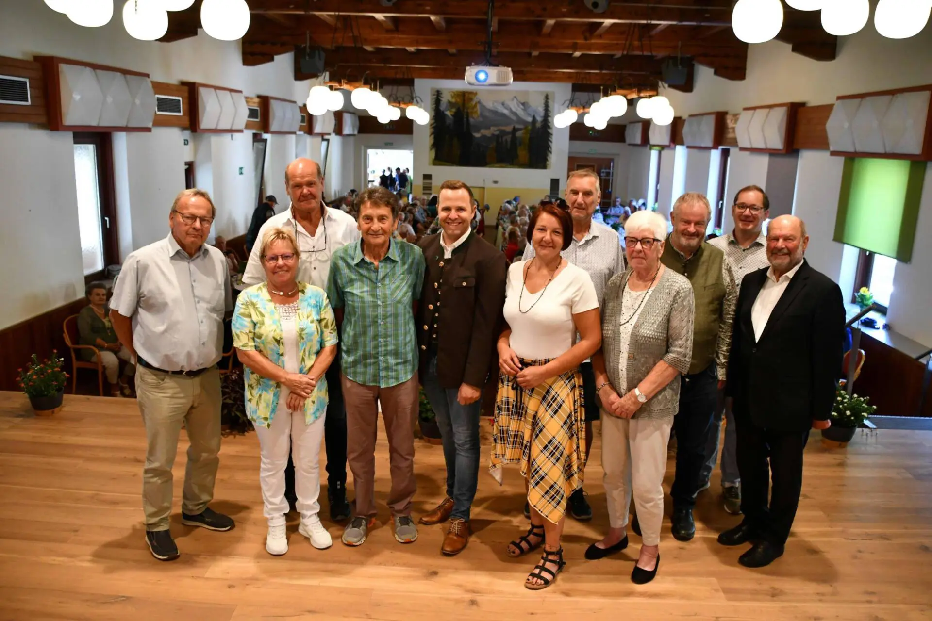 Eine Gruppe posiert für ein Foto in einem Saal mit Gästen im Hintergrund.