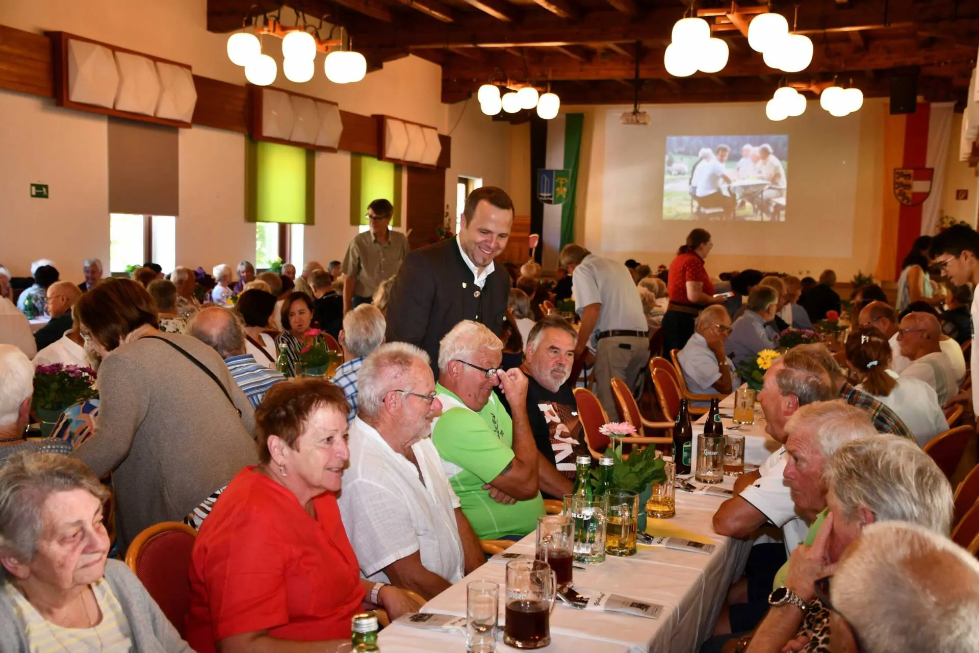 Gäste sitzen an gedeckten Tischen im Saal, während ein Mann zwischen ihnen steht.