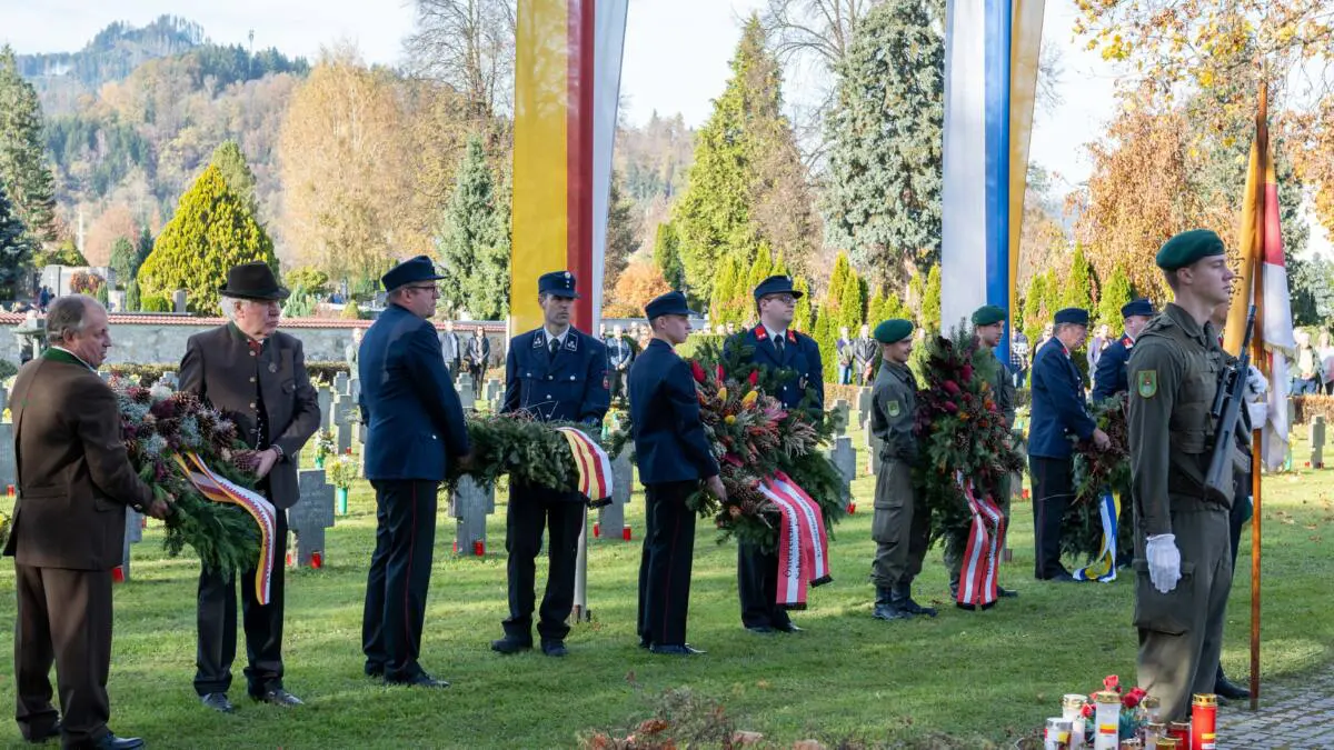 Uniformierte und Männer in Zivil halten Kränze auf einem Friedhof.