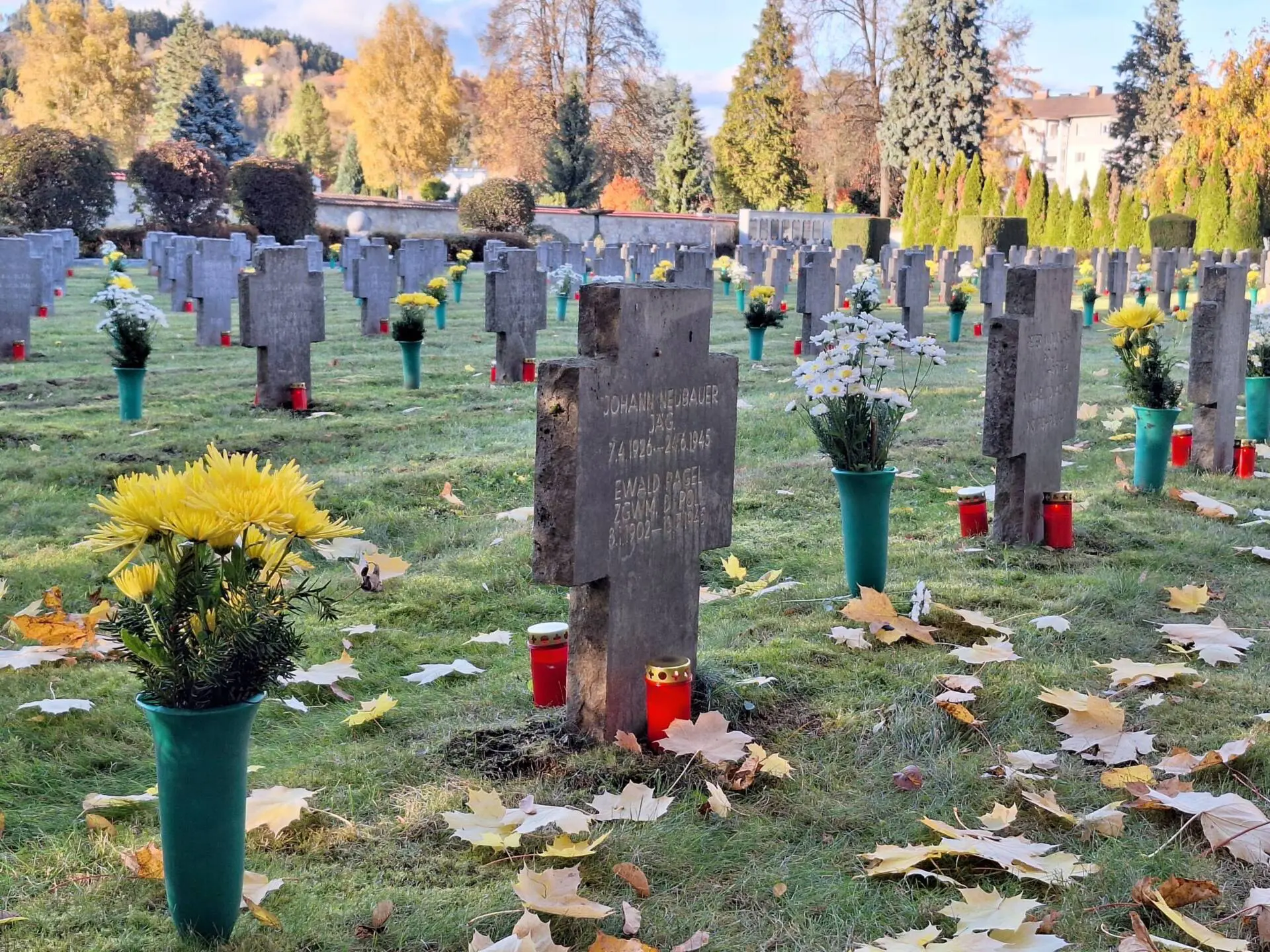 Steinerne Kreuzgrabmale mit Blumen und roten Grablichtern auf einem herbstlichen Friedhof.