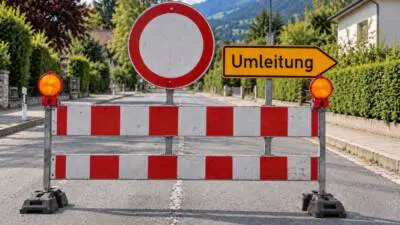 Rot-weiße Straßensperre mit gelbem Umleitungsschild auf einer Dorfstraße vor bewaldeten Bergen