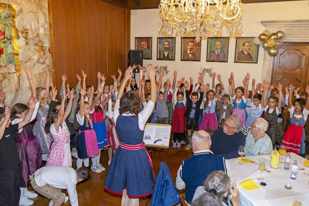 Kinder in Tracht heben bei einer Aufführung die Hände; Gäste sitzen an gedeckten Tischen.