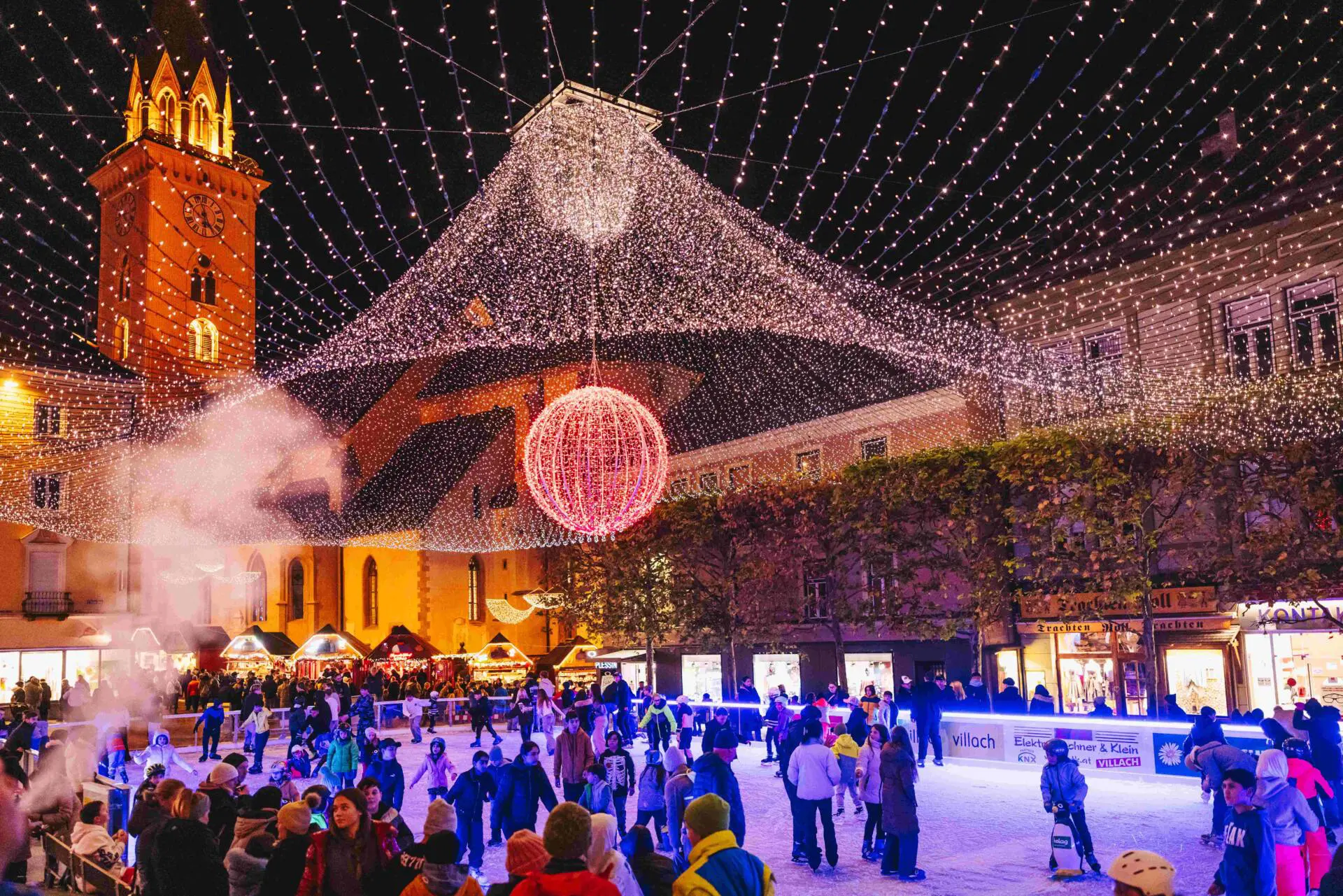 Besucher auf einer Eisfläche unter einem Lichterdach vor beleuchteten Gebäuden