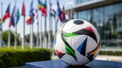 KI-Symbolbild: Ein Fußball mit bunten Mustern auf einem blauen Sockel vor Flaggen und einer Glasfassade.