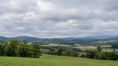 KI-Symbolbild: Graue Wolken über einer grünen Hügellandschaft mit Feldern.