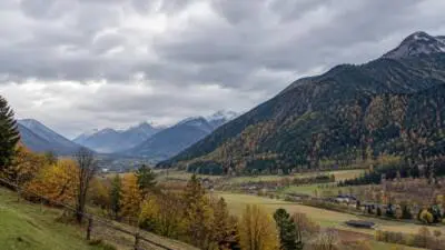 KI-generiertes Bild: Ein herbstliches Alpental mit bewaldeten Bergen unter dicht bewölktem Himmel.
