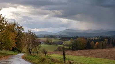 KI-generiertes Bild: Eine ländliche Hügellandschaft liegt unter dunklen Regenwolken; eine nasse Straße führt durch Felder.