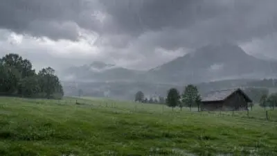KI-Symbolbild: Regen fällt auf eine Wiese vor Bergen und einer Holzhütte.