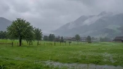 KI-Symbolbild: Regen über einer grünen Wiese mit Berglandschaft und Häusern.