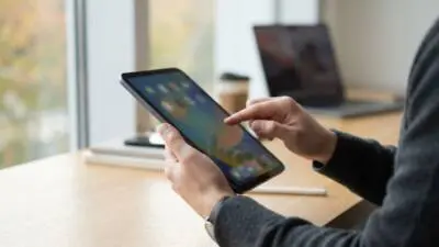 KI-Symbolbild: Eine Person tippt auf ein schwarzes Tablet an einem hellen Holztisch.