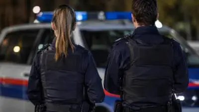 KI-generiertes Bild: Zwei Personen in dunkler Uniform stehen vor einem weiß-blauen Fahrzeug mit Blaulicht.