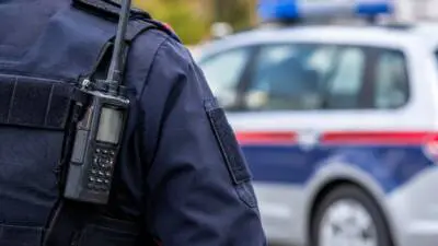 KI-Symbolbild: Eine dunkel gekleidete Person mit Funkgerät vor einem weiß-blau-roten Fahrzeug.