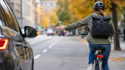 KI-Symbolbild: Eine Radfahrerin streckt neben einem dunklen Auto den Arm aus.