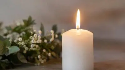 KI-Symbolbild: Eine brennende weiße Kerze steht neben kleinen weißen Blumen.