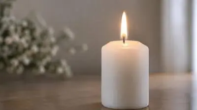 KI-Symbolbild: Eine brennende weiße Stumpenkerze steht auf einem Holztisch vor unscharfen Blüten.