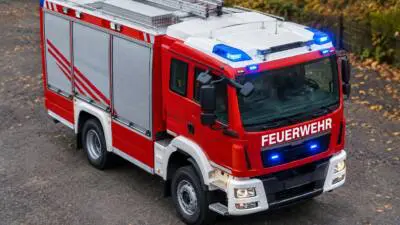 KI-generiertes Bild: Ein rotes Feuerwehrfahrzeug mit Blaulicht auf einer herbstlichen Fahrbahn.