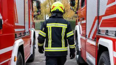 Eine Person in dunkler Schutzkleidung geht zwischen roten Einsatzfahrzeugen auf einer herbstlichen Straße.