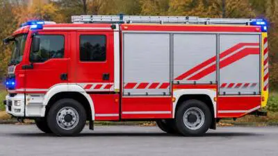 KI-generiertes Bild: Ein rotes Feuerwehrfahrzeug mit Dachleiter und blauem Blaulicht steht auf einer Straße.