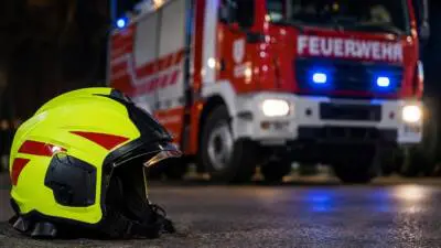 KI-generiertes Bild: Ein gelber Feuerwehrhelm liegt vor einem roten Feuerwehrfahrzeug mit eingeschaltetem Blaulicht.