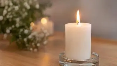 KI-generiertes Bild: Eine brennende weiße Kerze steht in einem Glasgefäß neben Blumen.