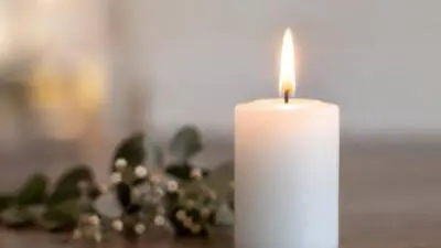 KI-generiertes Bild: Eine brennende weiße Kerze steht neben unscharfen Blumen auf einem Tisch.