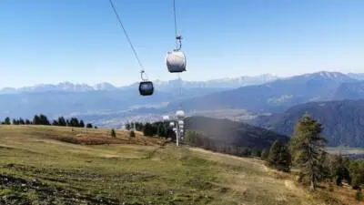 Seilbahngondeln über einer Bergwiese vor einem weiten Alpenpanorama