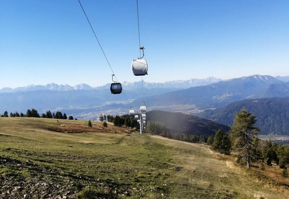Seilbahngondeln über einer Bergwiese vor einem weiten Alpenpanorama