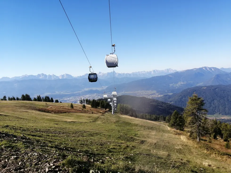 Seilbahngondeln über einer Bergwiese vor einem weiten Alpenpanorama