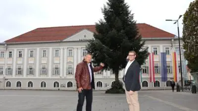 Zwei Männer stehen neben einem großen Nadelbaum auf einem Platz vor einem Gebäude.