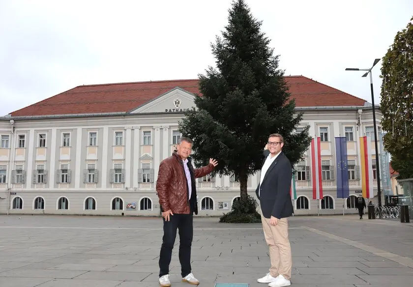 Zwei Männer stehen neben einem großen Nadelbaum auf einem Platz vor einem Gebäude.