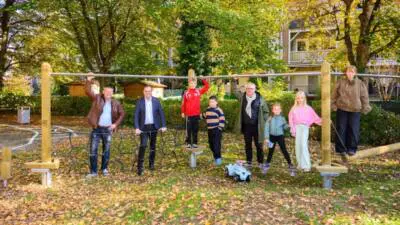 Kinder und Erwachsene stehen an einem hölzernen Spielgerät mit Querstrebe; daneben steht ein Bobbycar.