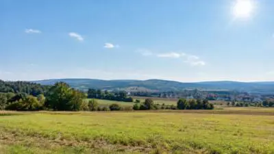 KI-Symbolbild: Sonnige Landschaft mit Feldern, Hügeln und einem Dorf unter blauem Himmel.