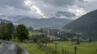 KI-Symbolbild: Eine nasse Straße führt durch ein grünes Bergtal unter dunklen Wolken.