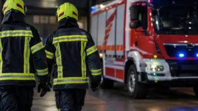 KI-generiertes Bild: Personen in dunkler Schutzkleidung und gelben Helmen vor einem roten Einsatzfahrzeug mit Blaulicht.