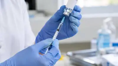 KI-generiertes Bild: Blaue Handschuhe halten ein Fläschchen und eine Spritze in einem hellen medizinischen Raum.
