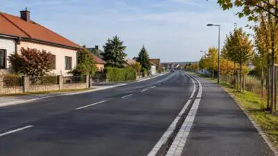 Eine asphaltierte Straße mit Gehweg verläuft zwischen Wohnhäusern und herbstlich gefärbten Bäumen.