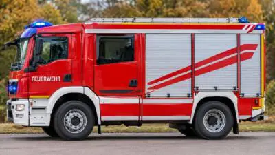 KI-generiertes Bild: Ein rotes Feuerwehrfahrzeug mit silbernen Geräteräumen und blauen Warnleuchten steht auf einer Straße.