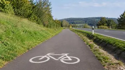 Asphaltierter Radweg mit weißem Fahrradsymbol verläuft neben einer Landstraße durch grüne Landschaft