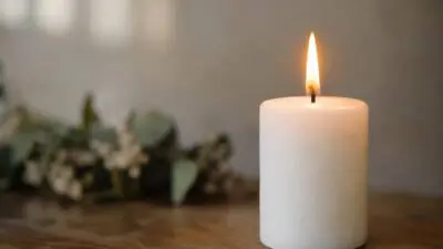 KI-generiertes Bild: Eine brennende weiße Kerze steht auf einem Holztisch vor unscharfen Blumen.