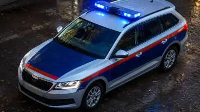 KI-Symbolbild: Ein silberner Kombi mit blau-roter Lackierung und eingeschaltetem Blaulicht auf nasser Straße.
