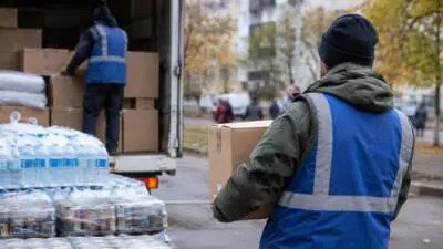 KI-Symbolbild: Eine Person in blauer Warnweste mit einem Karton vor einem Lastwagen mit Wasserflaschen und Kisten.