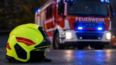 KI-generiertes Bild: Ein gelber Schutzhelm liegt vor einem roten Einsatzfahrzeug mit Blaulicht.