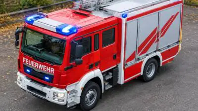 KI-Symbolbild: Ein rotes Feuerwehrfahrzeug mit Leiter und eingeschaltetem Blaulicht auf einer Straße.