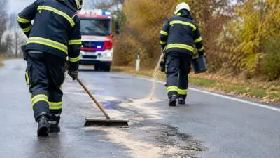 KI-generiertes Bild: Einsatzkräfte reinigen mit Besen und Eimer eine nasse Straße vor einem roten Einsatzfahrzeug.