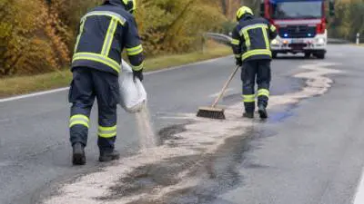 Personen in dunkler Schutzkleidung verteilen helles Bindemittel auf einer Straße.