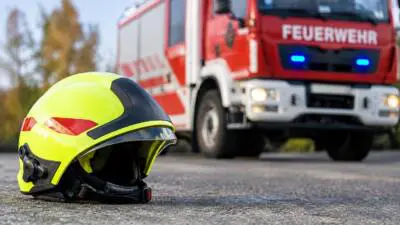 KI-generiertes Bild: Ein leuchtgelber Feuerwehrhelm liegt vor einem rot-weißen Feuerwehrfahrzeug mit blauem Blinklicht.