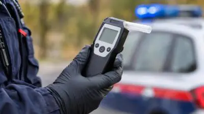 KI-Symbolbild: Eine behandschuhte Hand hält ein Atemalkoholmessgerät vor einem Fahrzeug mit Blaulicht.
